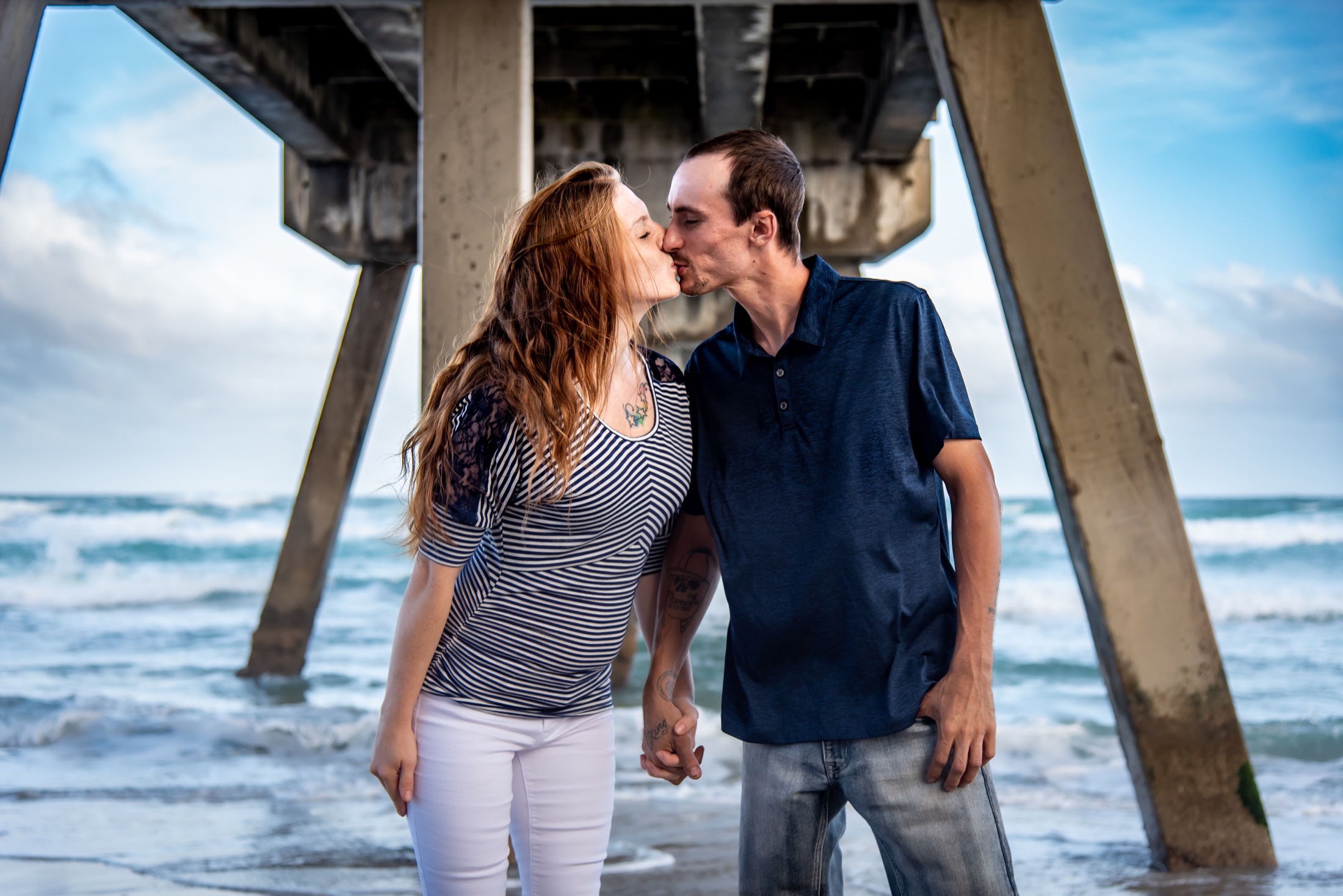 Expectant couple sharing a kiss beneath a South Florida beach pier