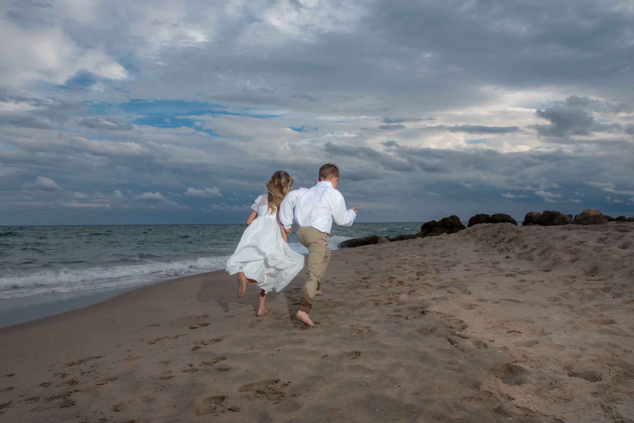 Siblings running along the shoreline in white and khaki