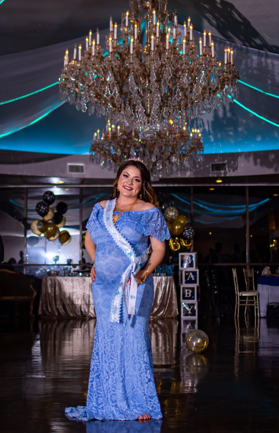 Expectant mother in a blue lace gown photographed beneath a dramatic chandelier